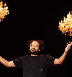 Photo Fabrice Eboué représentant l'humoriste les bras levés et deux luminaires accrochés au plafond à Le Mée-sur-Seine